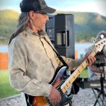 A man wearing a cap is playing a bass guitar outdoors. He is in front of a keyboard setup and mountains are visible in the background.