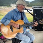 A man with a gray beard and ponytail, wearing a cap and denim shirt, plays an acoustic guitar and sings into a microphone outdoors, with distant mountains and greenery in the background.