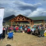 A crowd sits on lawn chairs facing a wooden stage at an outdoor event. An Estes Park Health banner stands on the left. Mountains and cloudy skies are in the background.