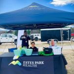 Three people sit under a blue Estes Park Health tent at an outdoor event. The table displays promotional items and two baskets, one with a sign marked "For Donations." Various vehicles are in the background.