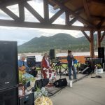 A music band performs under a wooden pavilion with mountains in the background. One person plays guitar, another is seated with a microphone, and a third stands singing into a microphone.