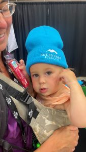 A child wearing a blue hat and an orange dress is being carried by an adult in a baby carrier. The hat has "Estes Park Health" written on it.