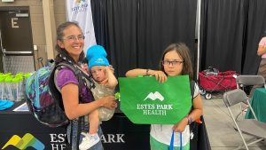 A woman holding a baby and a child holding a green bag stand in front of a display table at an Estes Park Health event.