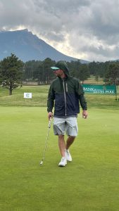 A person in a green jacket and shorts holds a golf club while standing on a green at a golf course, with mountains and trees in the background.