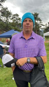 A man wearing a blue beanie and a patterned polo shirt stands on a grassy field, holding a black jacket and a white hat. Trees and an event tent are visible in the background.