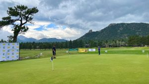 Two people stand on a green golf course with various sponsor banners in the background, surrounded by trees and mountains under a cloudy sky.