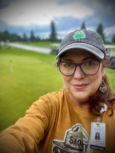 A person wearing glasses and a cap, with a name badge, stands outdoors on a grassy area.