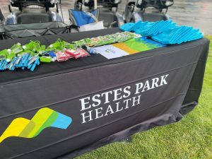 A table with a black tablecloth displaying the "Estes Park Health" logo, laid out with various promotional items such as pens, hand sanitizers, and blue gloves.