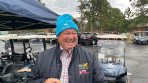 A man wearing a blue beanie and a dark jacket is smiling while standing in a parking lot filled with golf carts. Trees and a cloudy sky are visible in the background.