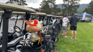 A row of golf carts lined up with various golf clubs and bags. Two people stand nearby, one looking inside a cart. Pine trees and mountains are visible in the background under a cloudy sky.