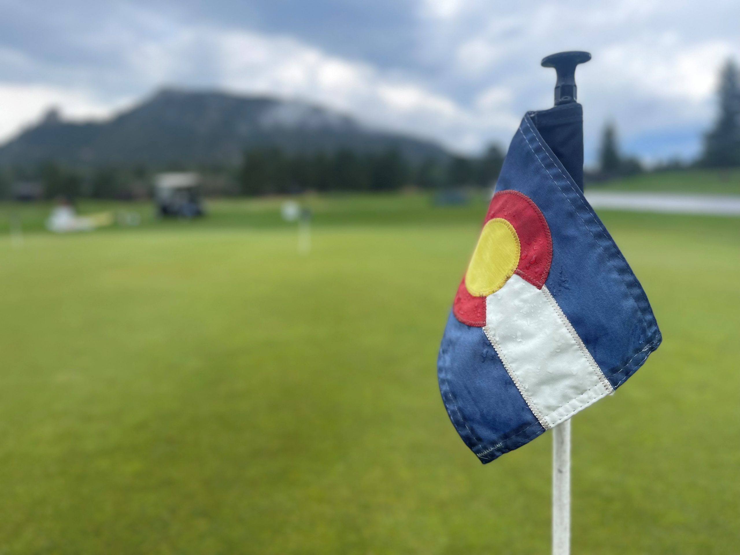 A golf hole flag featuring the Colorado state flag design stands on a green with a mountain and cloudy sky in the background.