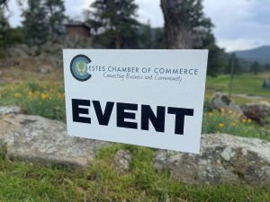 A close-up of a sign reading "EVENT" hosted by Estes Chamber of Commerce, placed outdoors on a grassy area with rocks and trees in the background.