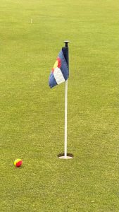 A golf hole with a small flag displaying the Colorado state emblem, and a yellow and red golf ball lying near the hole on a green grassy field.