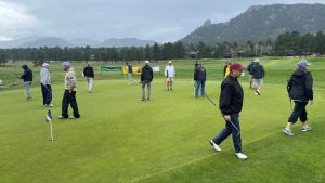 A group of people, dressed in casual sportswear, stand on a golf course green with a mountainous backdrop and overcast sky. Some hold golf clubs while others converse.