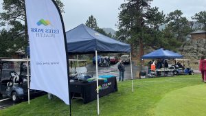 Outdoor event featuring tents, including one labeled "Estes Park Health," with tables and people setting up or browsing in a parking lot area on a rainy day. Golf carts and a grassy area are visible.