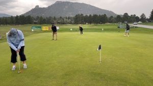 Golfers practice putting on a green with mountains and trees in the background. Four people are visible, each focusing on their own putt. The sky is overcast.