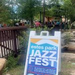 A sign promotes the Estes Park Jazz Fest. In the background, people are sitting and standing at an outdoor venue surrounded by trees.