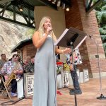 A woman in a grey jumpsuit sings into a microphone on an outdoor stage with musicians seated behind her.