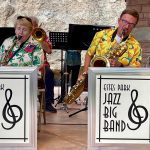 Two saxophone players, one man and one woman, perform with the Estes Park Jazz Big Band. They are seated with music stands displaying the band's name and are wearing floral shirts.