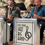 Musicians from a jazz big band perform in front of a rocky backdrop. The band name "Estes Park Jazz Big Band" is displayed on the music stands. Saxophones and trombones are prominent.