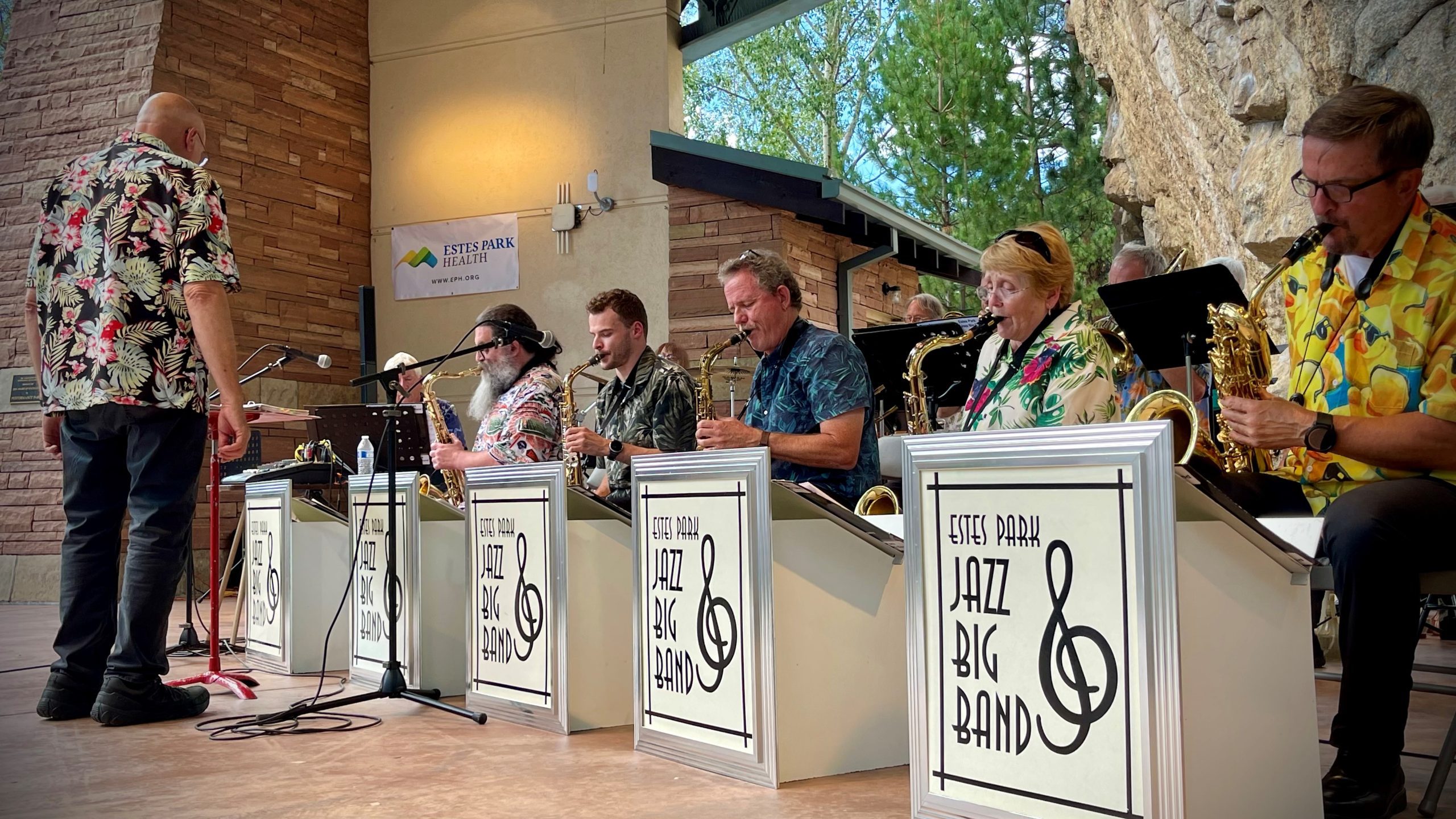 A jazz big band performs on stage with saxophones, trumpets, and a conductor, all wearing colorful shirts. The stage backdrop includes brick walls and a sign reading “Estes Park Jazz Big Band.”.