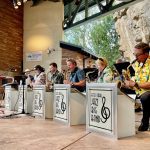 A jazz big band performs on an outdoor stage, with six musicians seated and one standing. The stage features rocky and brick walls, and the musicians wear colorful shirts.