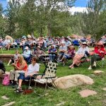 A large group of people sit outdoors in folding chairs on a grassy slope, attending an event. Many are wearing hats and sunglasses. Trees and greenery surround them.