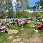 A large group of people sit on folding chairs and blankets on a grassy area surrounded by trees, attending an outdoor event.