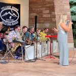 A woman in a grey dress sings into a microphone on an outdoor stage while accompanied by a jazz big band, consisting of musicians playing various instruments such as saxophones and trumpets.