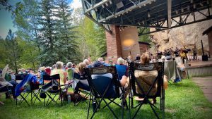 Audience sitting on lawn chairs watching an outdoor concert at a park with a stage and musicians. Trees and rocky cliffs are visible in the background.