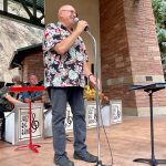 A man in a floral shirt speaks into a microphone on an outdoor stage with a jazz band setup behind him.