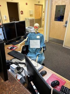 A Halloween skeleton prop in hospital scrubs sits at a computer desk with a sign that reads, "Epic - Go Live! I'm ready." The scene is in an office with medical equipment and multiple monitors.