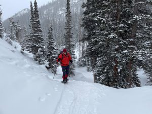 A person dressed in red winter gear is snowshoeing on a trail through a snowy forested mountain landscape.