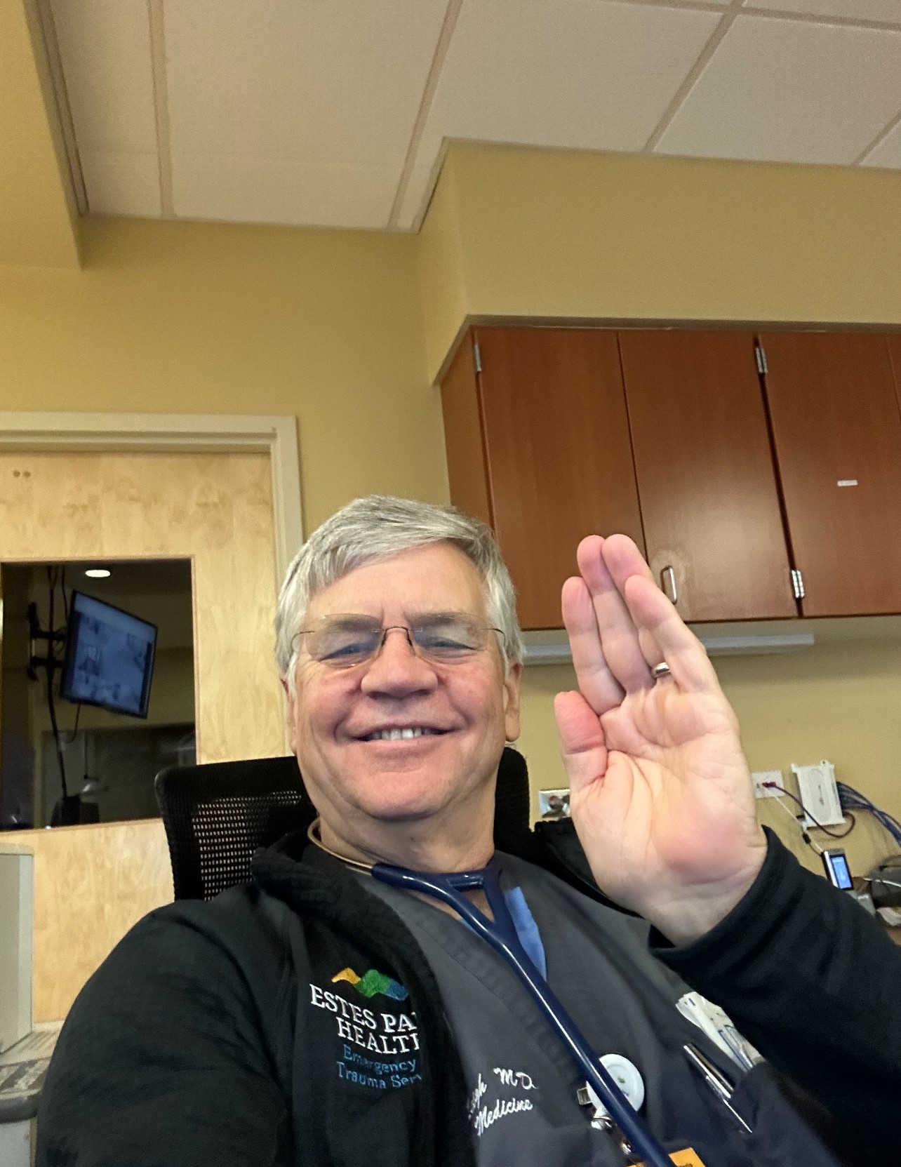 A person with glasses and gray hair wearing a medical uniform is seated indoors, smiling and raising their hand in a waving gesture. An office environment with wooden cabinets and a computer monitor is visible.