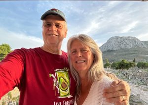 A man in a red shirt and cap stands next to a woman with long gray hair in an outdoor setting with ancient ruins and a mountain in the background.