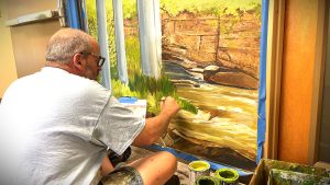 A person in glasses is painting a landscape mural of a river and trees on a wall indoors. Several paint containers are on the floor nearby.