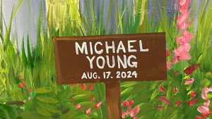 A wooden sign in a garden reads "Michael Young, Aug. 17, 2024" written in white paint. The background features tall green plants and pink flowers.