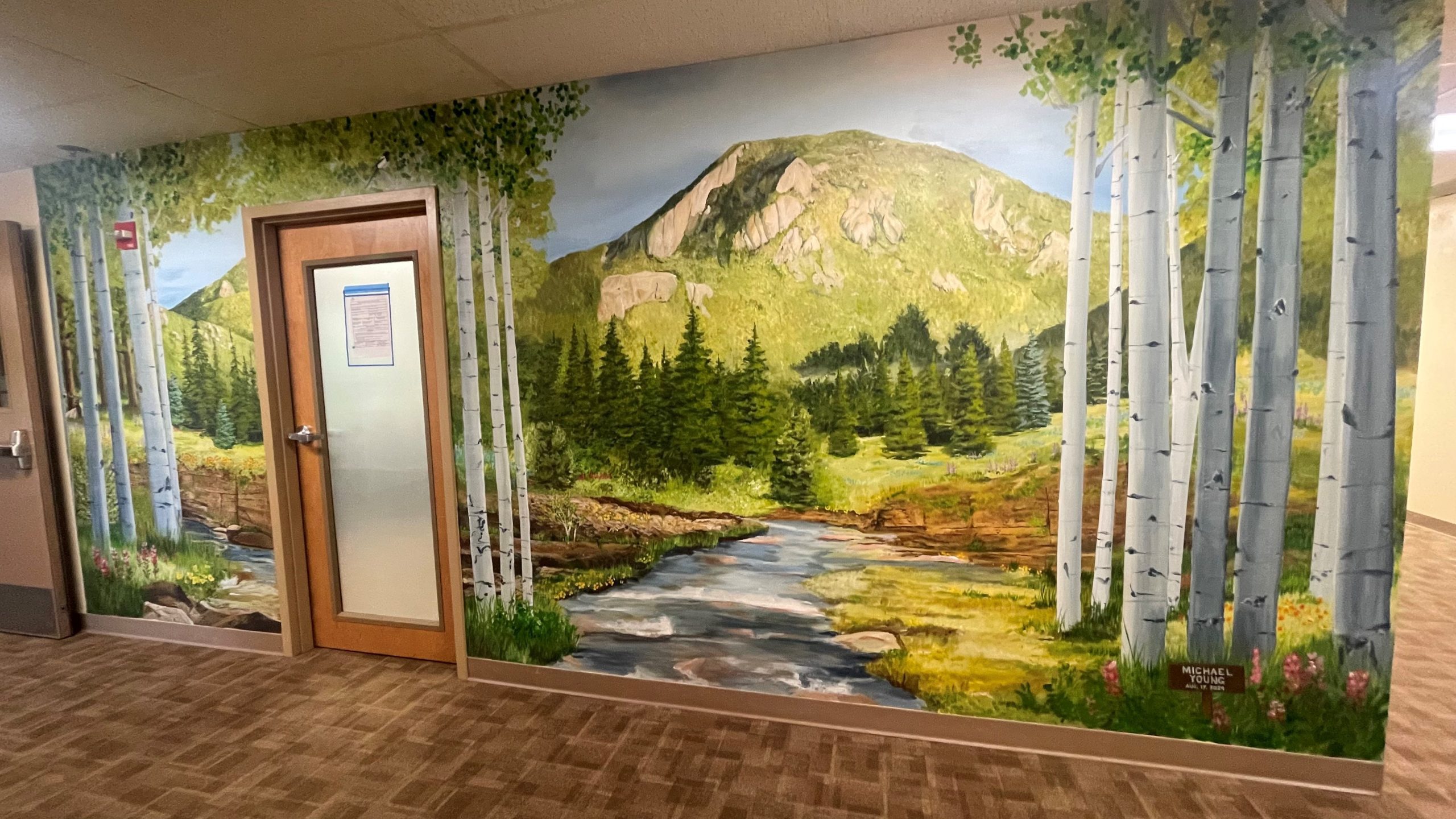 A hallway wall mural depicting a scenic landscape with birch trees, a flowing stream, greenery, and a mountain in the background. A closed door on the left has a notice posted on it.