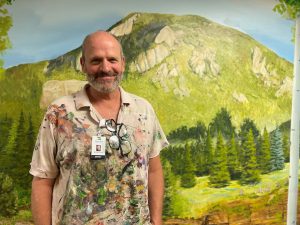 A man stands in front of a painted mural depicting a green mountainous landscape. He wears a paint-stained shirt and an ID badge with glasses clipped to it.