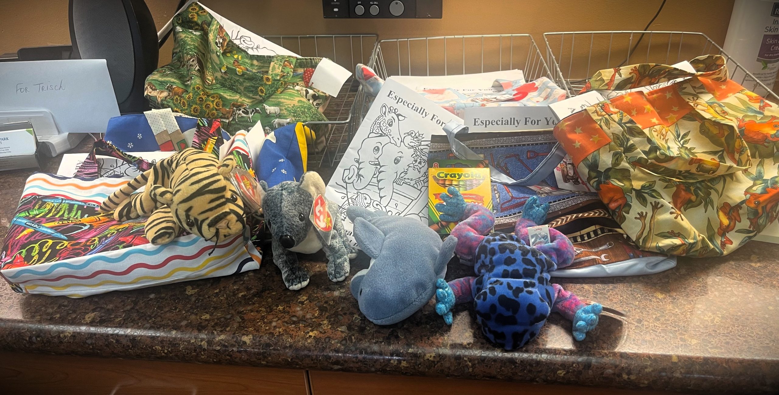 A cluttered desk surface with various items including stuffed animals, a crayon box, assorted fabric, and coloring pages.