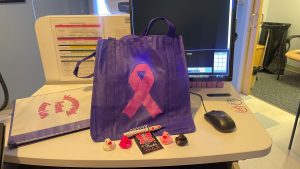 A desk with a computer, a mouse, a blue bag with a pink ribbon, several rubber ducks, a pen, and a binder with documents.