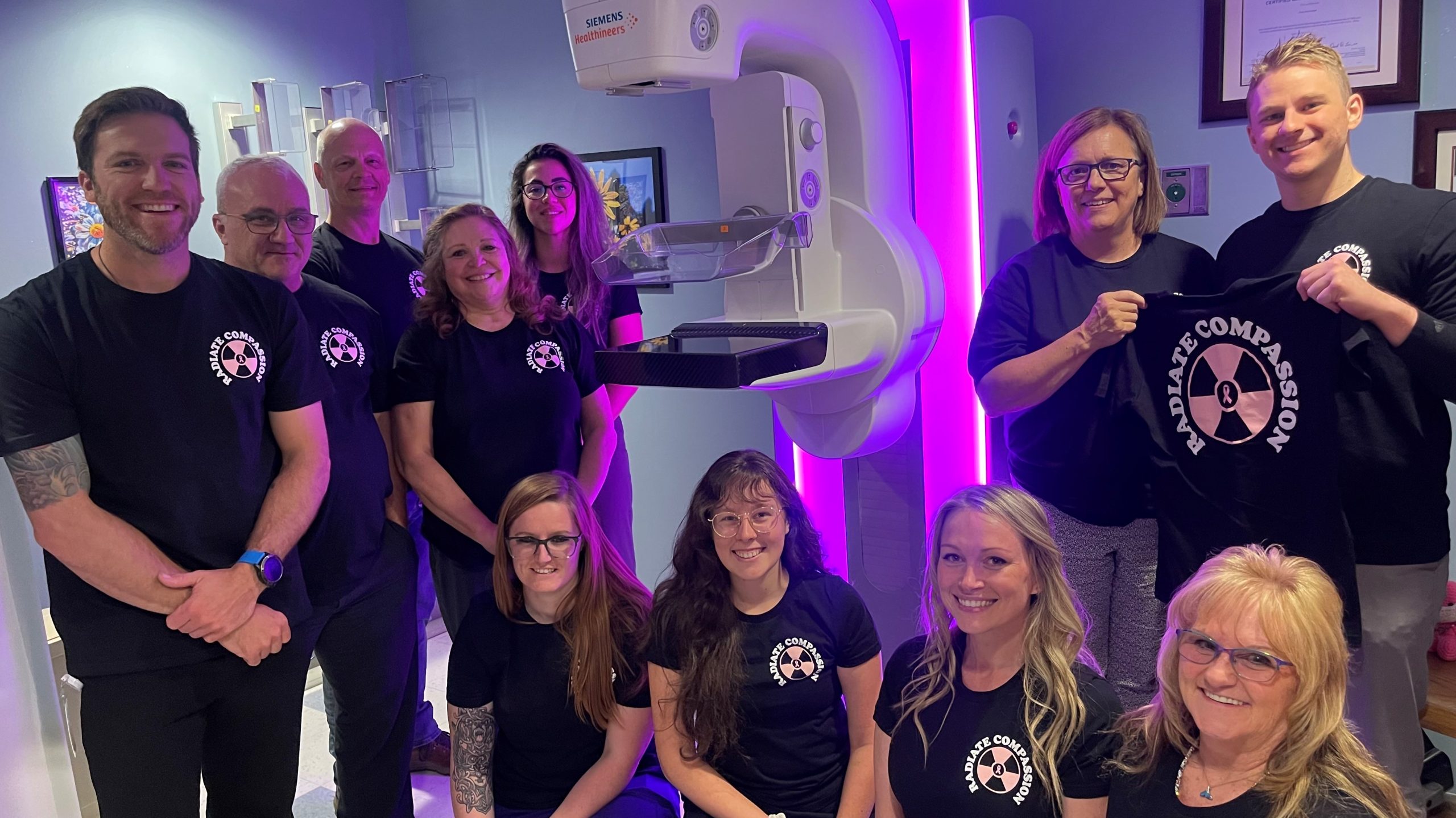 Group of ten people wearing matching shirts posing by a mammography machine, with two individuals holding a shirt that reads 