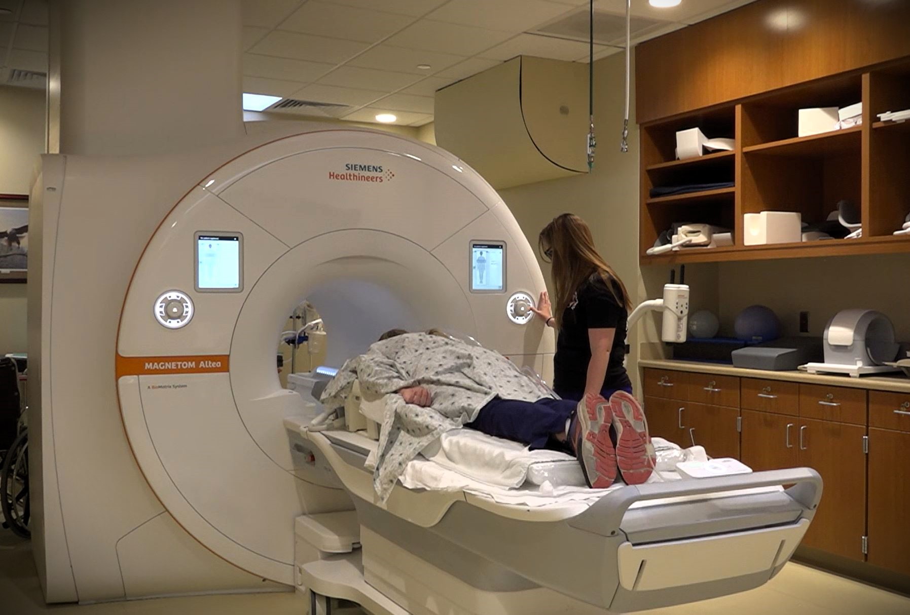 A person lying on a bed is being prepared to enter a large MRI machine by a medical professional in a clinical setting. Various medical equipment and storage cabinets are visible in the room.