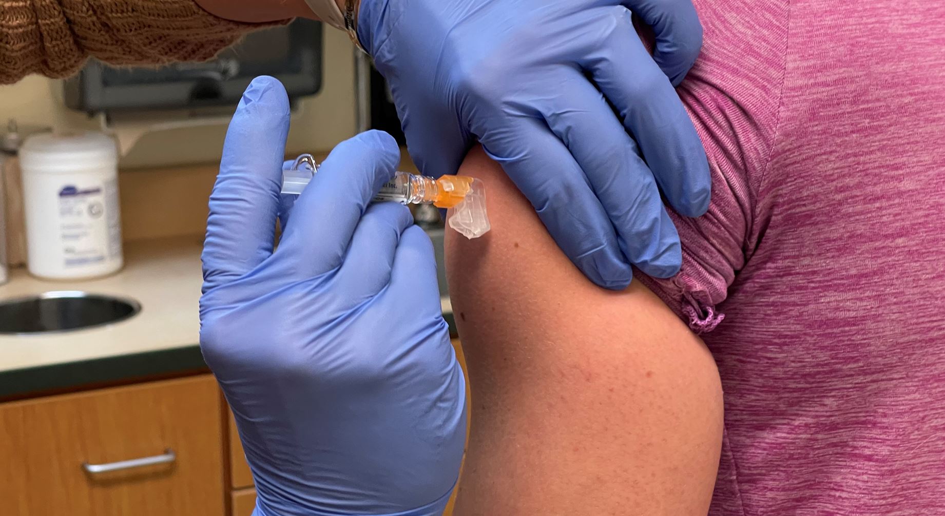 A person in a pink shirt receives an injection in their arm from a medical professional wearing blue gloves.