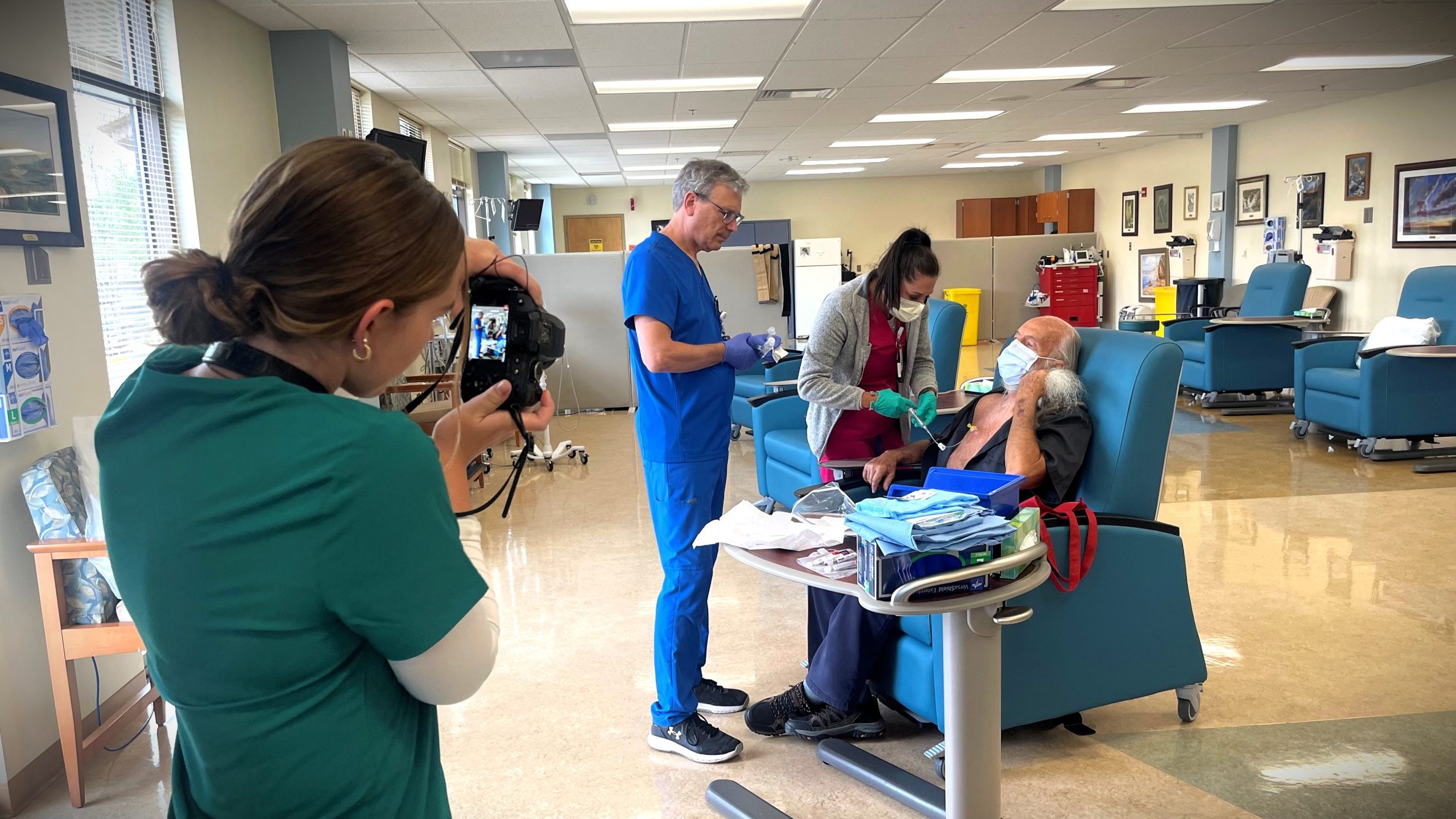 Three healthcare workers in a clinical setting attend to an elderly patient seated in a medical chair. One nurse is taking photos, while another adjusts medical equipment. The patient is holding an oxygen mask.