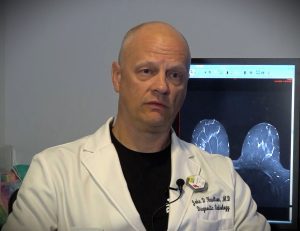 A bald man in a white lab coat labeled "diagnostic radiology" sits in front of a medical imaging screen showing scans.