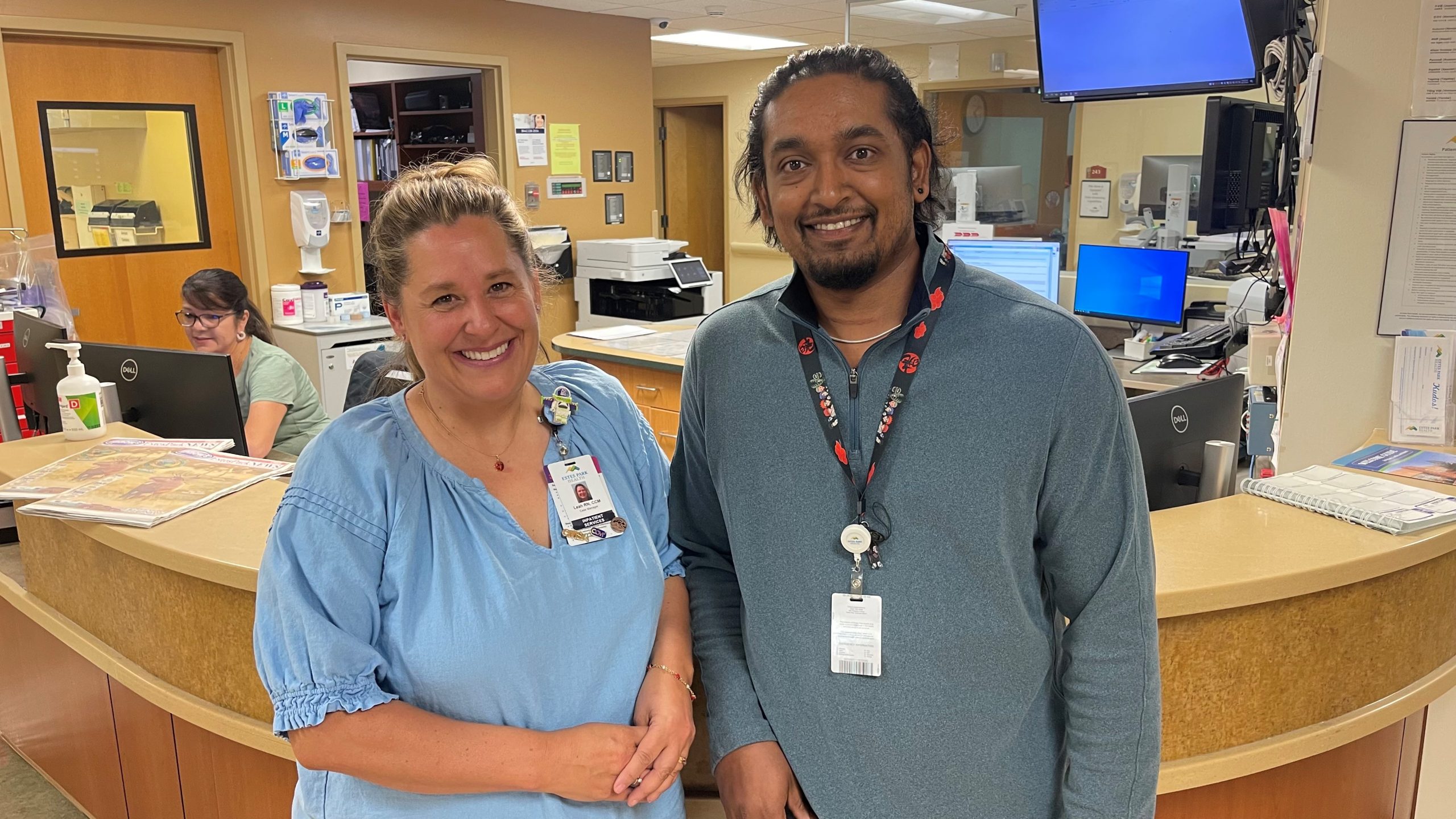 Two people standing and smiling in a hospital workspace, with computers and office supplies in the background. Both are wearing ID badges.