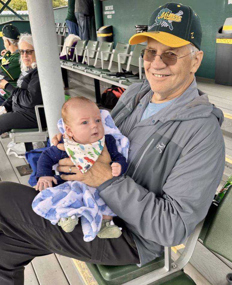 An older man wearing glasses and a baseball cap holds a baby wrapped in a blue blanket at an outdoor stadium.