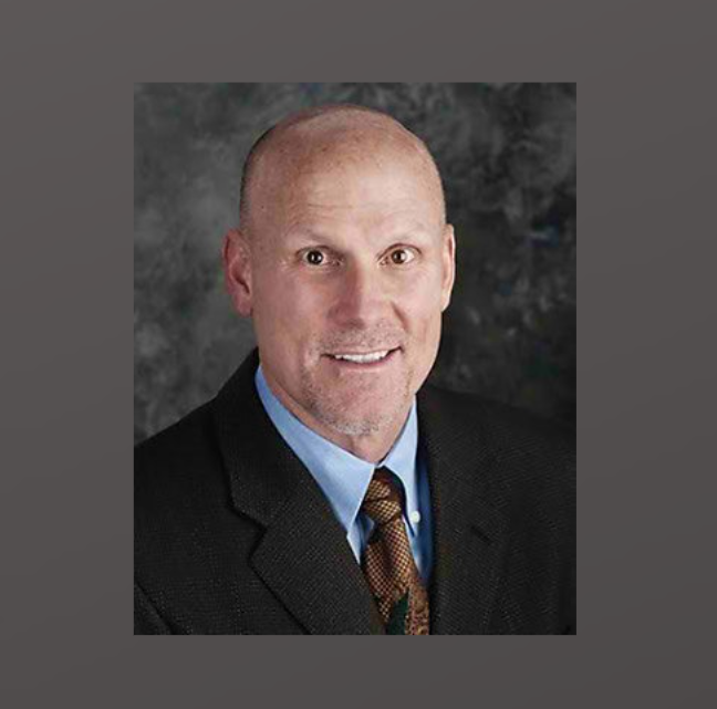 A bald man in a dark suit, light blue shirt, and tie, smiles in a professional headshot with a dark, textured background.