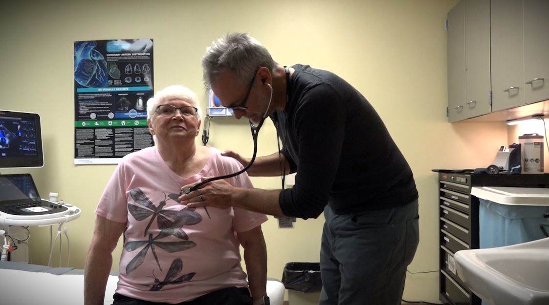 A doctor uses a stethoscope to examine the heart of a seated woman in a medical office.